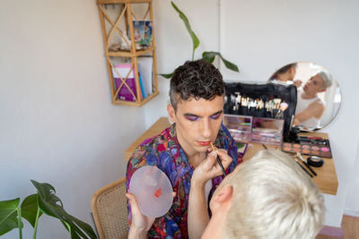 Young androgynous man getting makeup done by his boyfriend at dressing table