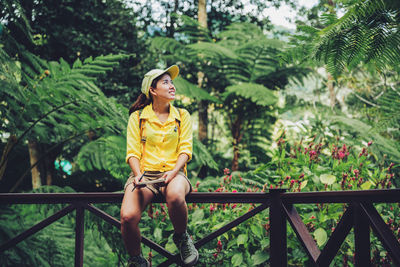 Young woman looking away while sitting on railing against trees