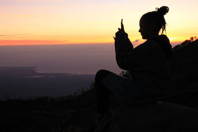 Silhouette man sitting on mountain against orange sky