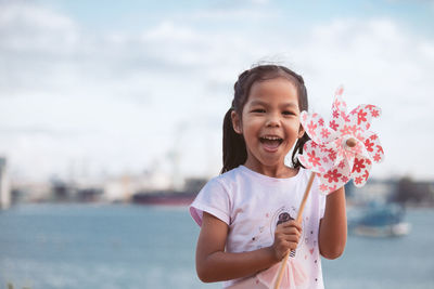 Portrait of cheerful girl holding pinwheel toy at beach against sky