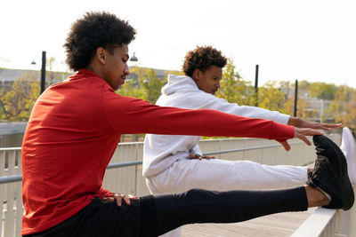 Side view of african american male doing hamstring stretch with leg on street fence near black friend