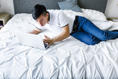 High angle of focused latin teen boy with down syndrome lying on bed and surfing internet on netbook