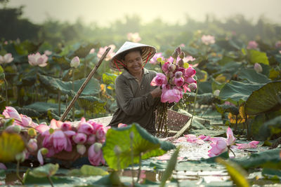 Smiling female farmer wearing asian style conical hat collecting lotus water lilies from pond in boat