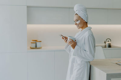 Smiling young woman wearing eye patches while holding mobile phone and coffee cup at home