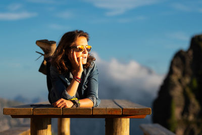 Young woman wearing sunglasses sitting on table against sky