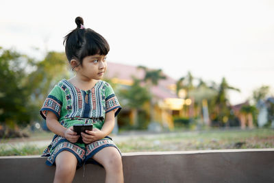 Girl using phone while sitting on retaining wall against sky