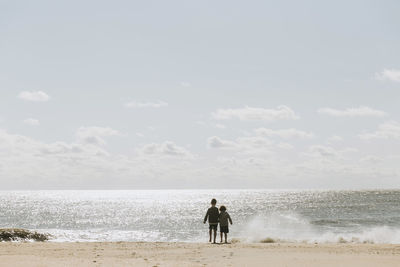 Rear view of brothers standing at shore against sky during sunny day