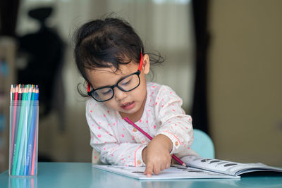 Little girl wearing glasses drawing on book at table.