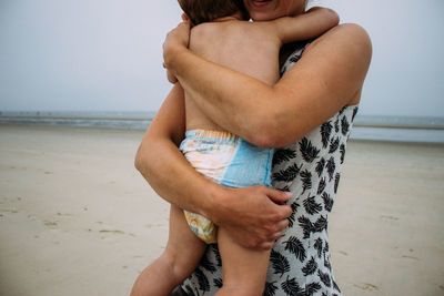 Midsection of woman holding her baby and standing at beach against sky.