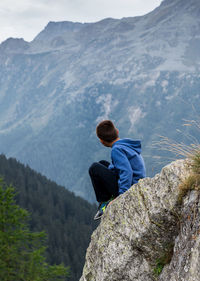 Side view of boy sitting on cliff by mountain range