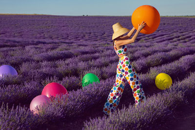 Fashionable young woman in hat on lavender field stands with colored balls and clothes to the point