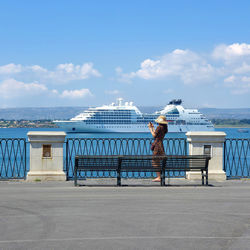 Side view of woman photographing with smart phone while standing on footpath by sea against sky during sunny day