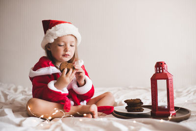 Cute child girl 3-4 year old wear red santa claus hat and bathrobe sit in bed with biscuit 