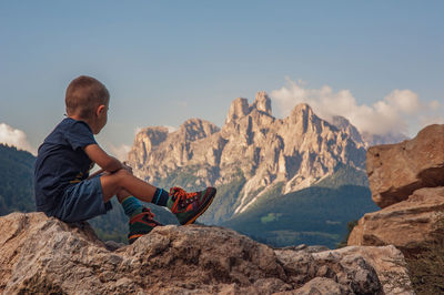 Side view of boy sitting on rock against mountains