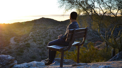 Man sitting on bench against mountains during sunset