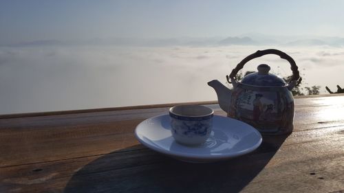 Cup and saucer by teapot on table against sky
