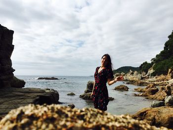 Portrait of young woman standing on rocky shore against cloudy sky