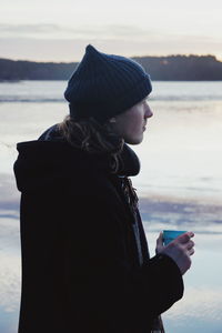 Side view of man having coffee while standing against lake during sunset