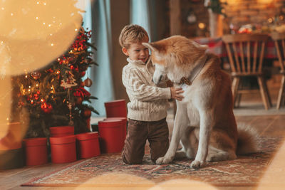 Candid authentic happy little boy in knitted beige sweater hugs dog with bow tie at home on xmas