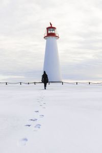 Rear view of person walking on snow covered field towards lighthouse against cloudy sky