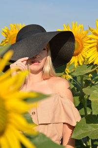 Beautiful woman standing wearing hat amidst sunflower plant against blue sky