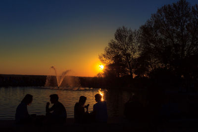 Silhouette people sitting on shore against sky during sunset