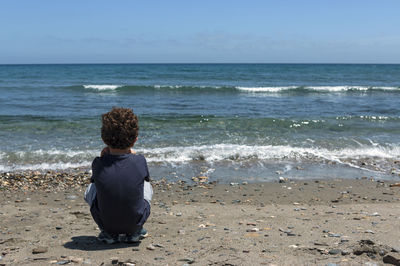 Little boy,  with brown and curly hair, alone on the beach watching the sea in a squatting position.