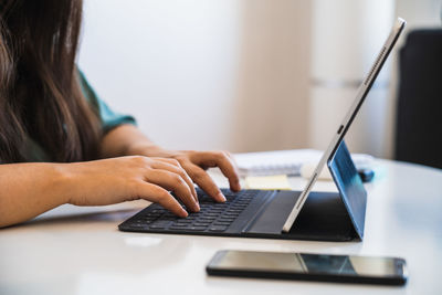 Close up of a woman's hands typing on the keyboard of her hybrid tablet and laptop device next to a phone on a table, while teleworking and adapting to the new normal after coronavirus crisis