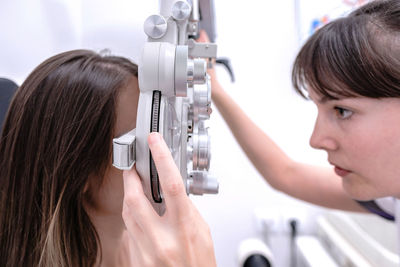 Side view of focused female optometrist using phoropter and checking eyesight of woman during check up in modern clinic