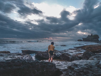 Rear view of man standing on rock by sea against cloudy sky