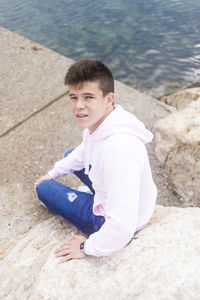 High angle portrait of young man sitting on rock at beach