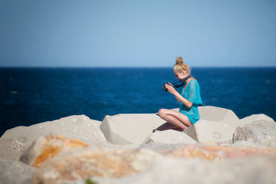 Side view of young woman looking at camera while sitting on rocks by sea