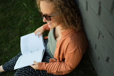 Young adult millennial with curly hair smiles and sits on grass. student reads book. lifestyle