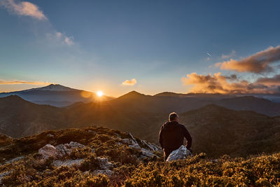 Man sitting on top of the cliff in mountains at sunset enjoying sunset over erupting volcano etna. 