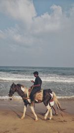 Side view of man riding horse at beach against cloudy sky