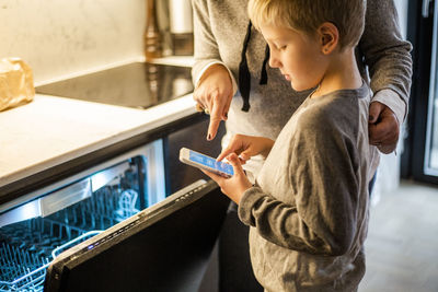 Boy learning from mother while using mobile app over dishwasher in kitchen at smart home