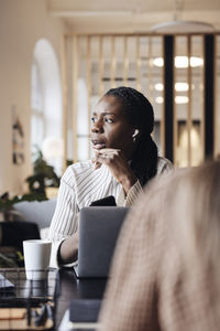 Thoughtful businesswoman looking away while talking through wireless in-ear headphones at coworking office