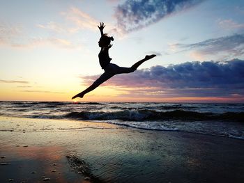 Young woman jumping on shore at beach against sky during sunset