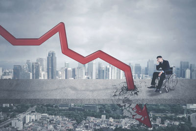 Businessman sitting on cracked footpath with graph against illuminated cityscape