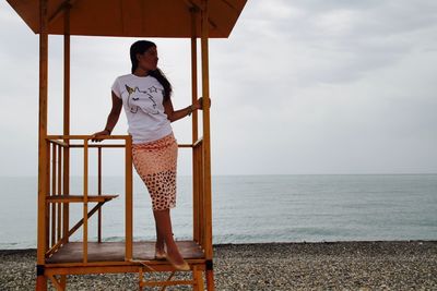 Full length of young woman standing in lifeguard hut at beach against cloudy sky