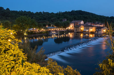 Illuminated buildings by lake against sky at night