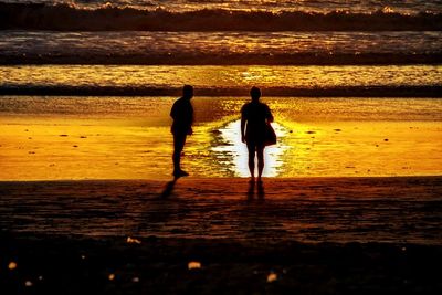 Rear view of silhouette friends standing at beach during sunset