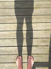 Low section of woman standing by shadow on boardwalk during sunny day