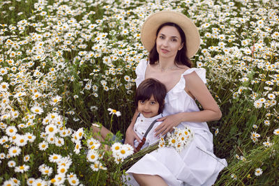 Woman in a white dress with her son, lies in a chamomile field at sunset in summer