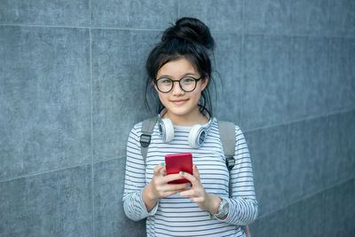 Portrait of teenage girl holding smart phone while standing against wall