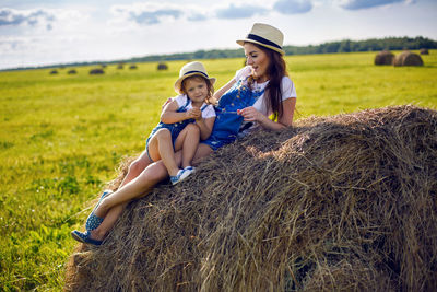 Daughter with mom standing in the field at the sheaf in hats and denim jumpsuits during sunset