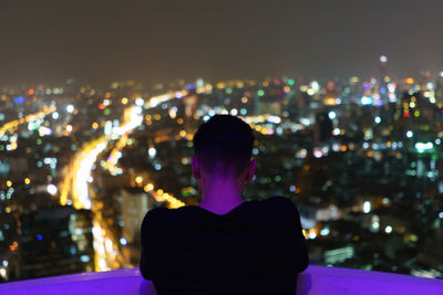 Rear view of woman sitting against illuminated cityscape at night