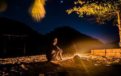 Man sitting by bonfire against sky at night