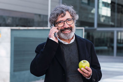 Happy senior businessman calling with mobile phone while eating a apple outside the office
