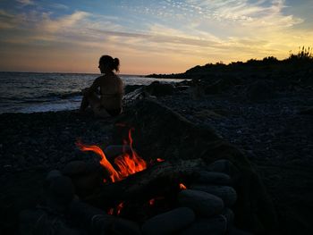 Man sitting on rock at beach against sky during sunset